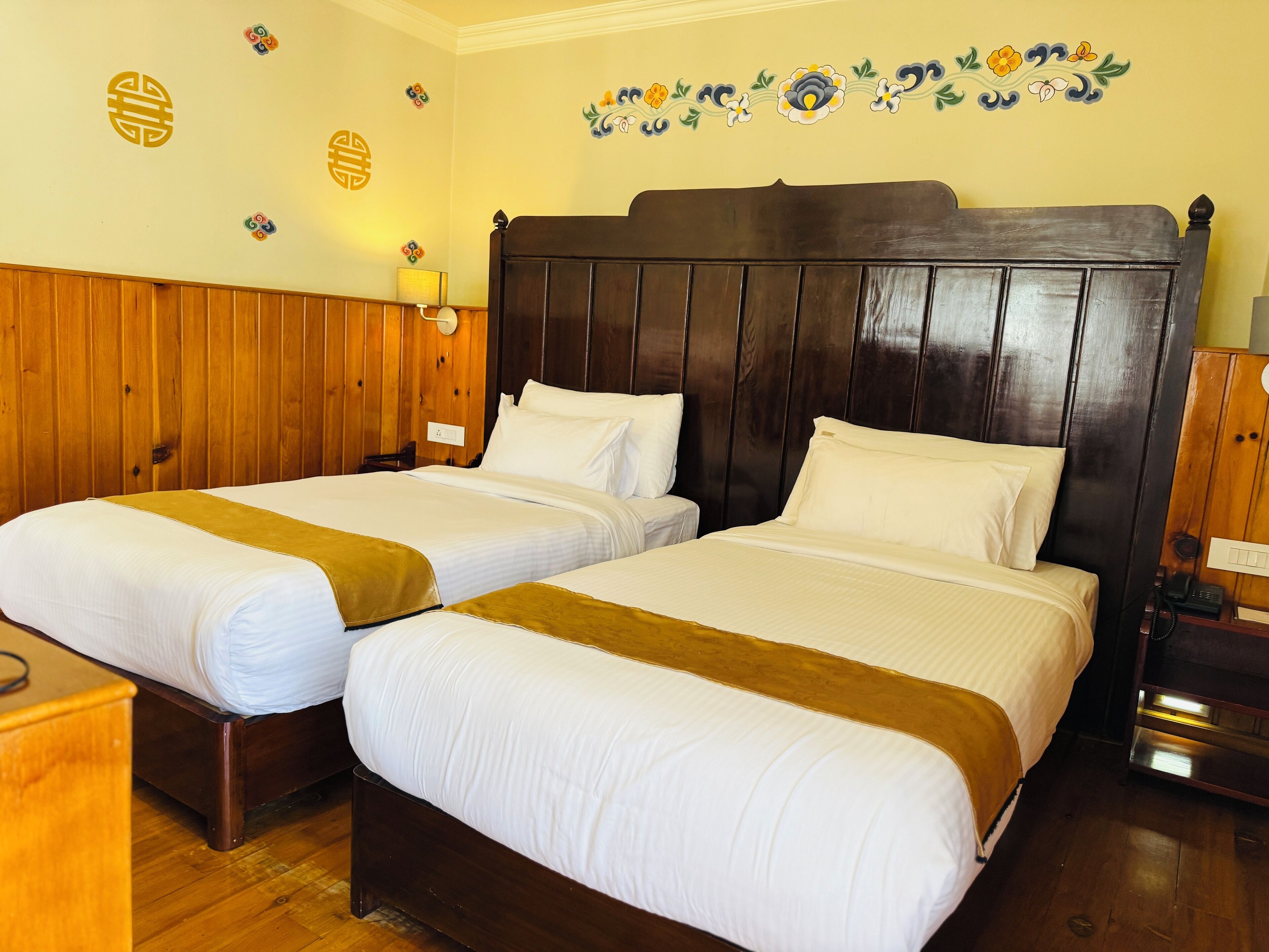 Une chambre d’hôtel avec deux lits en bois, une tête de lit en bois et une décoration murale à motifs floraux.