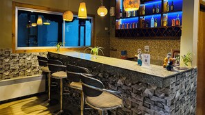 Zona de bar con pared de piedra, taburetes, luces colgantes y estantes con botellas.