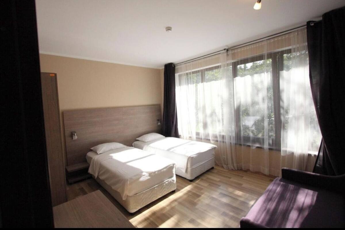Ein Hotelzimmer mit zwei Betten, einem großen Fenster mit durchsichtigen Vorhängen und Holzboden.