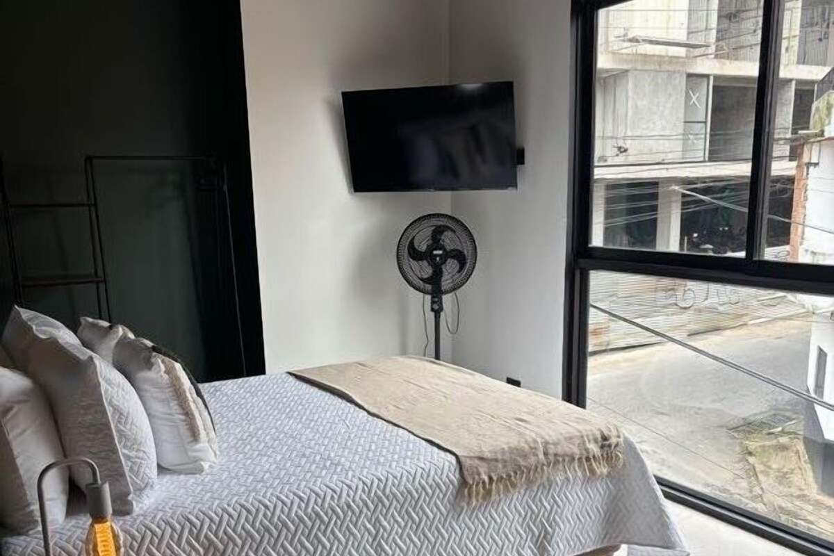 Un dormitorio moderno con una cama, un televisor, un ventilador y un amplio ventanal.