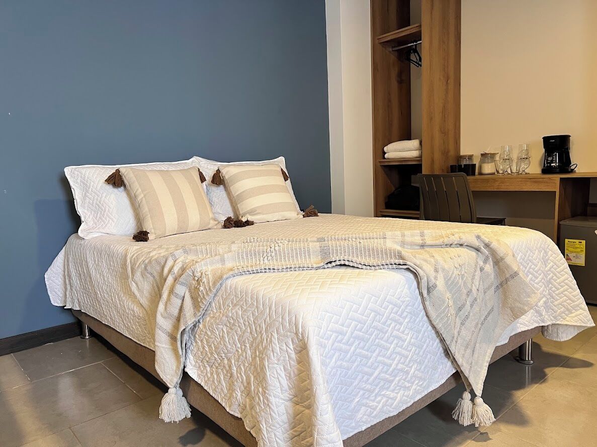 Un letto rifatto con copriletto trapuntato e cuscini, un comodino in legno con una macchina per il caffè e un mobile contenitore con asciugamani piegati e bicchieri.