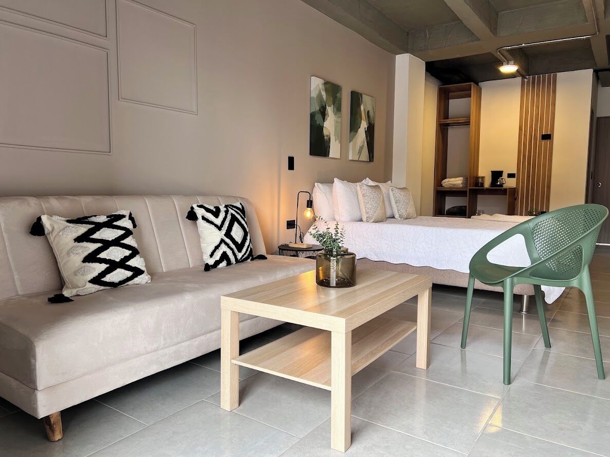 A modern hotel room with a sofa, a wooden coffee table, a green chair, a bed, and abstract wall art.