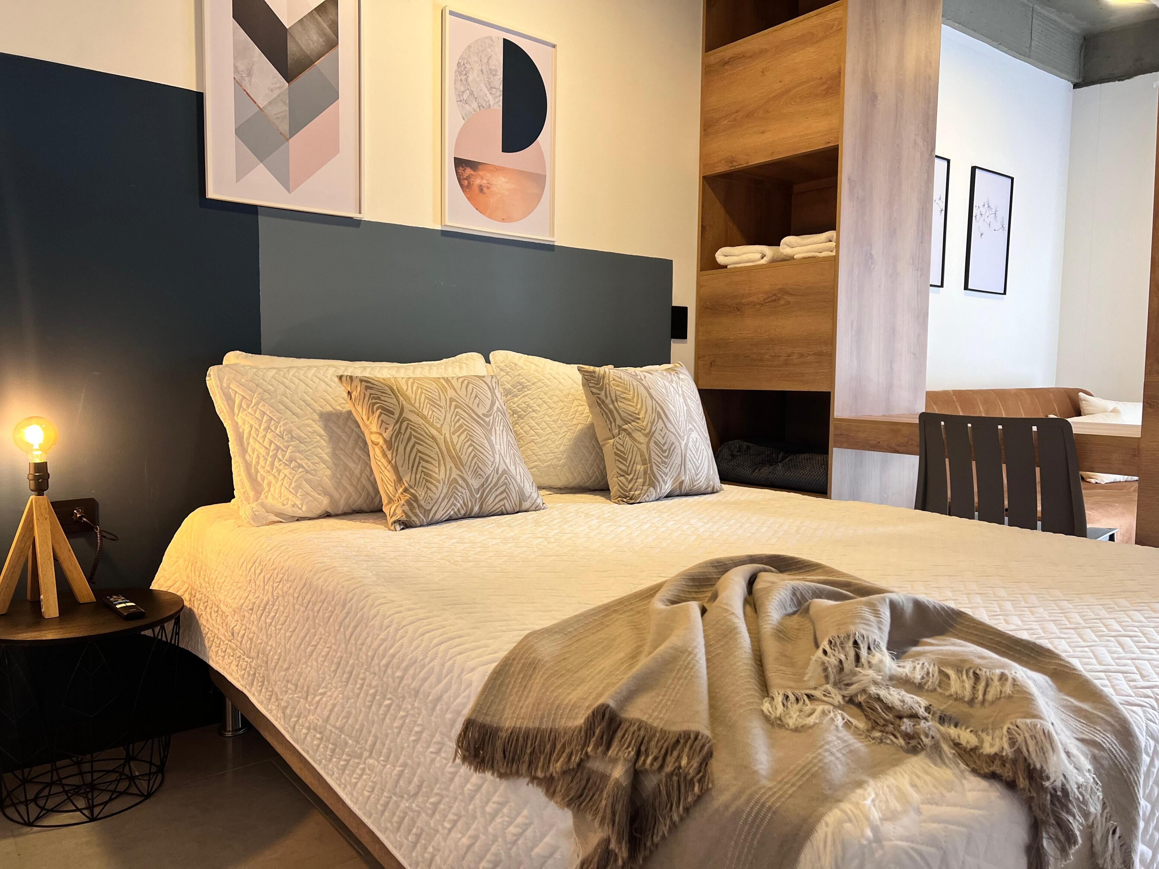A neatly made bed with pillows, a bedside lamp, and a blanket. There are framed artworks on the wall and a wooden shelf with towels.