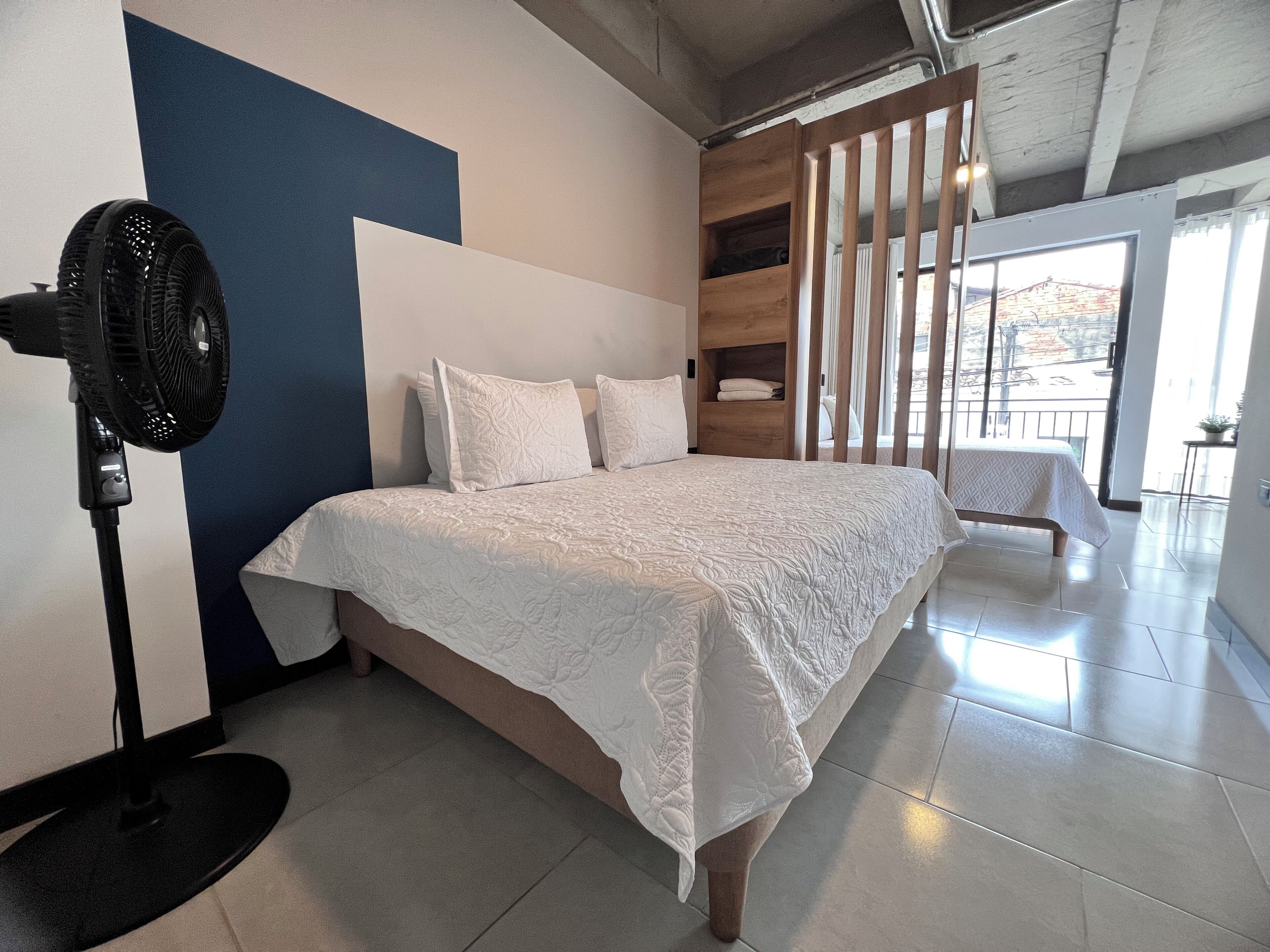 Un dormitorio con cama, un ventilador de pie, un estante de madera y vistas al exterior.