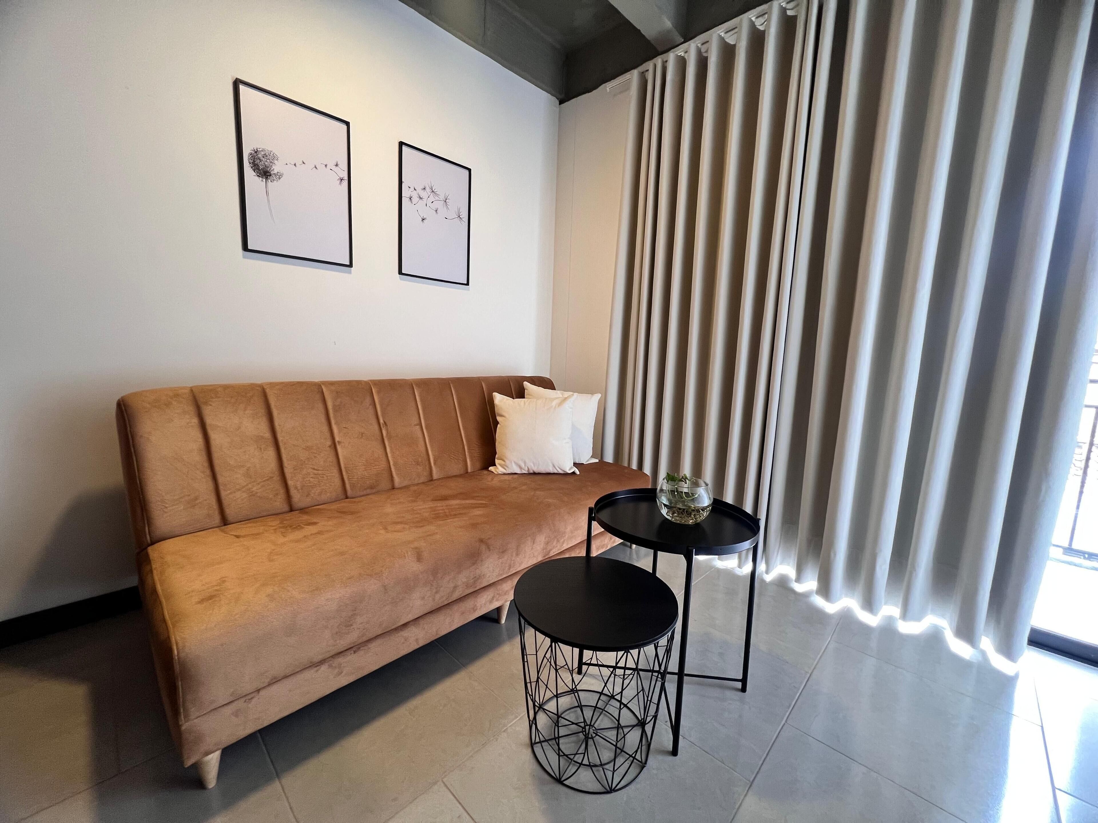A modern living room with a brown sofa, two framed artworks, a round black table with a wireframe base, and a large window with vertical blinds.