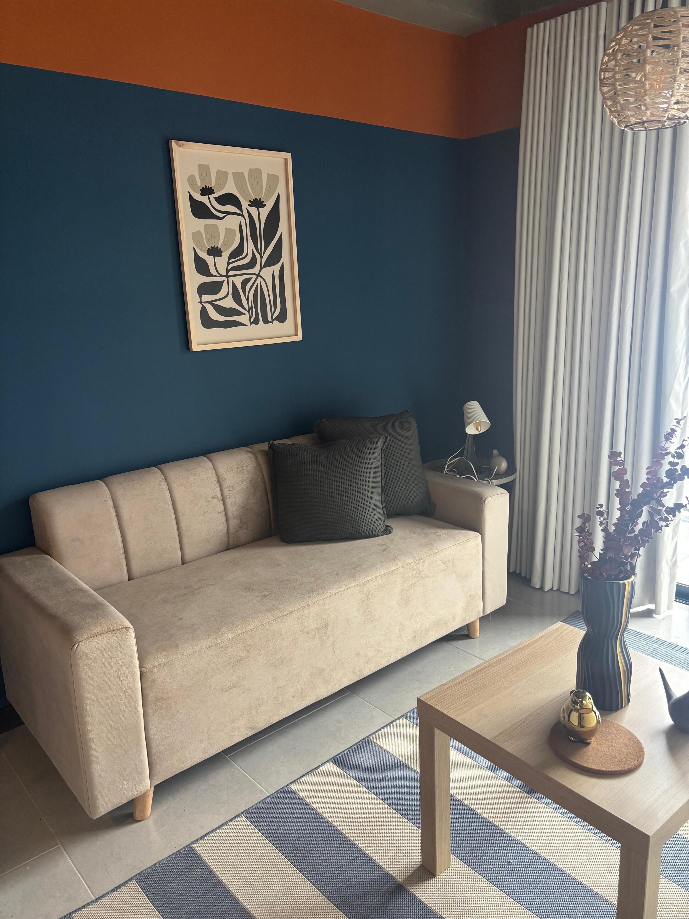 A living room with a beige sofa, a coffee table, a lamp, a vase with flowers, and a framed abstract artwork on the wall.