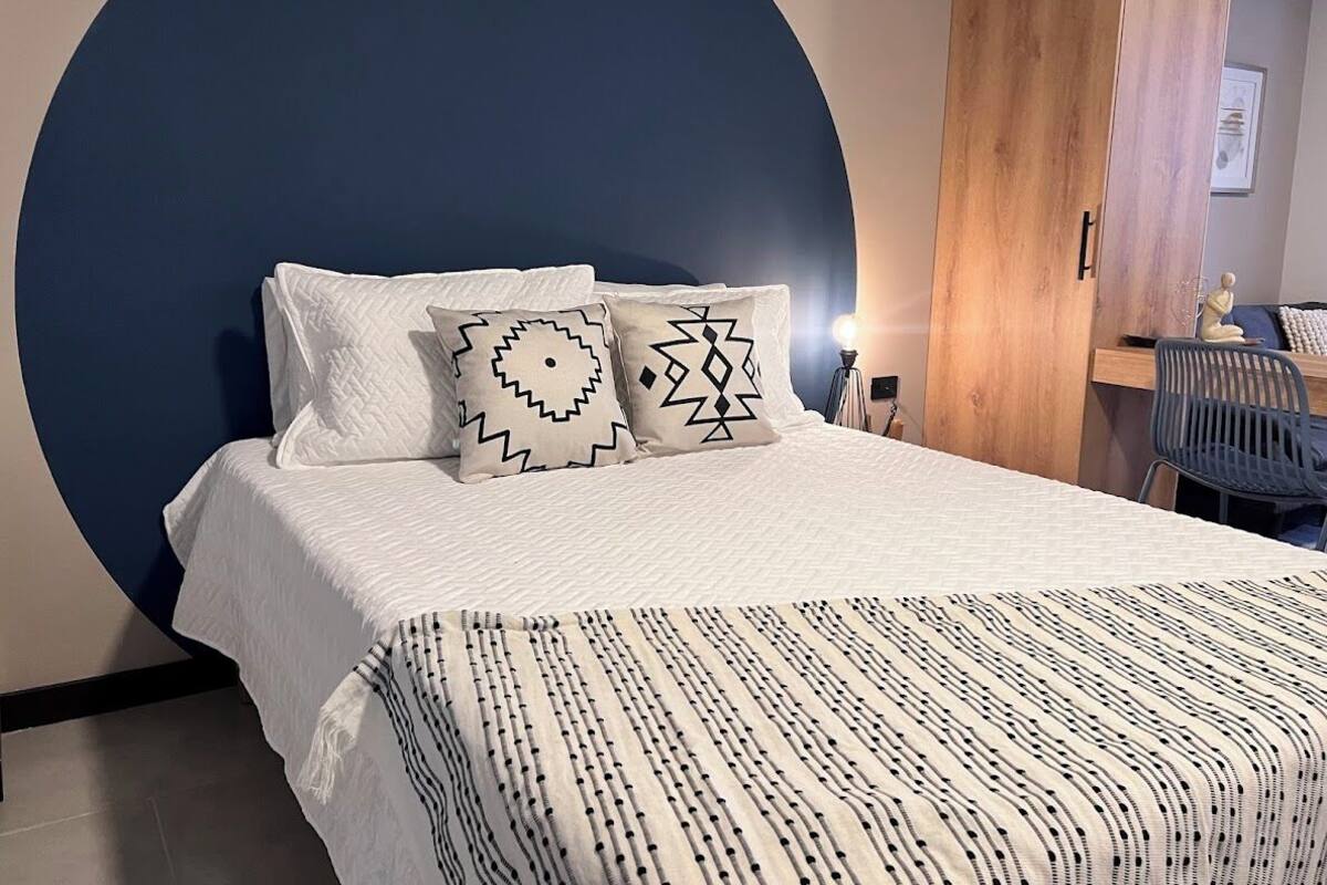 A bed with a white bedspread and two patterned pillows, a wooden door, and a chair in a room with a large circular wall feature.