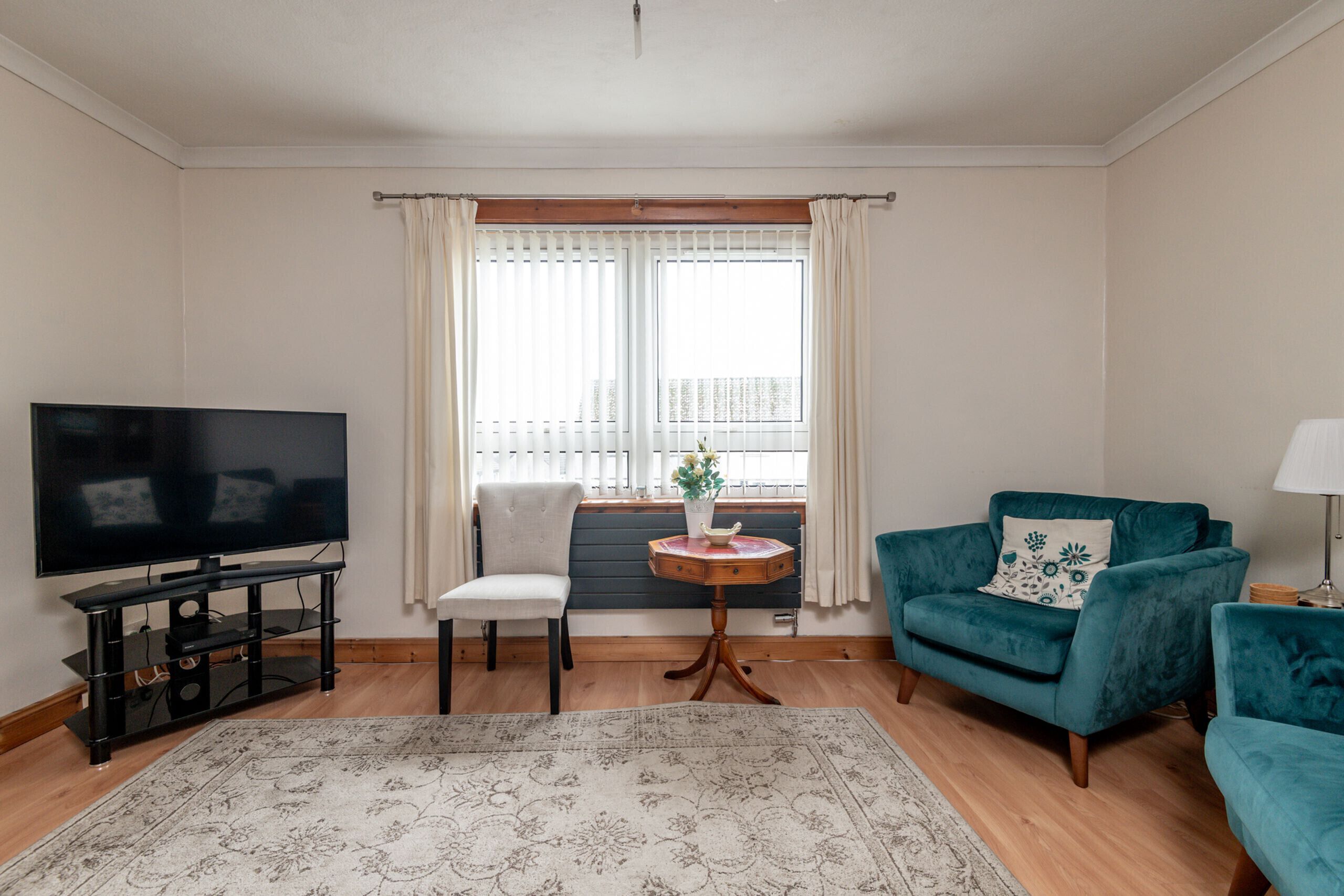 A living room with a flat-screen TV, a teal armchair, a white chair, a small round table with a vase, and a large patterned rug.