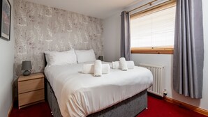 Una cama bien tendida con sábanas y almohadas blancas, una mesita de noche con lámpara y una ventana con cortinas grises.