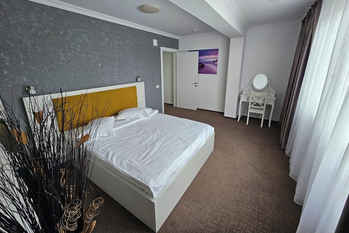 A modern hotel room with a large bed, a desk with a mirror, and a door leading to another room.
