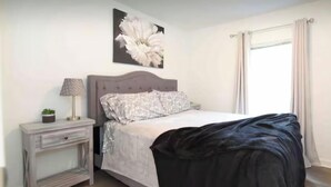 A bedroom with a bed, a nightstand, a lamp, a wall-mounted flower painting, and a window with curtains. - Cozy Bungalow Retreat (Des Moines)