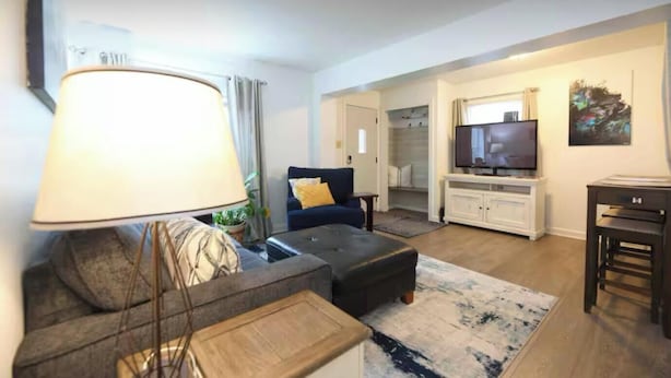 A living room with a sofa, ottoman, lamp, TV, and a painting on the wall. - Cozy Bungalow Retreat (Des Moines)