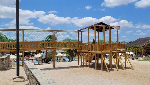 Una estructura de juego de madera con un área cercada, una piscina y un entorno de playa.