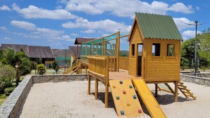Una estructura de madera para juegos con un tobogán y una red de escalada, rodeada por un muro de piedra y follaje.