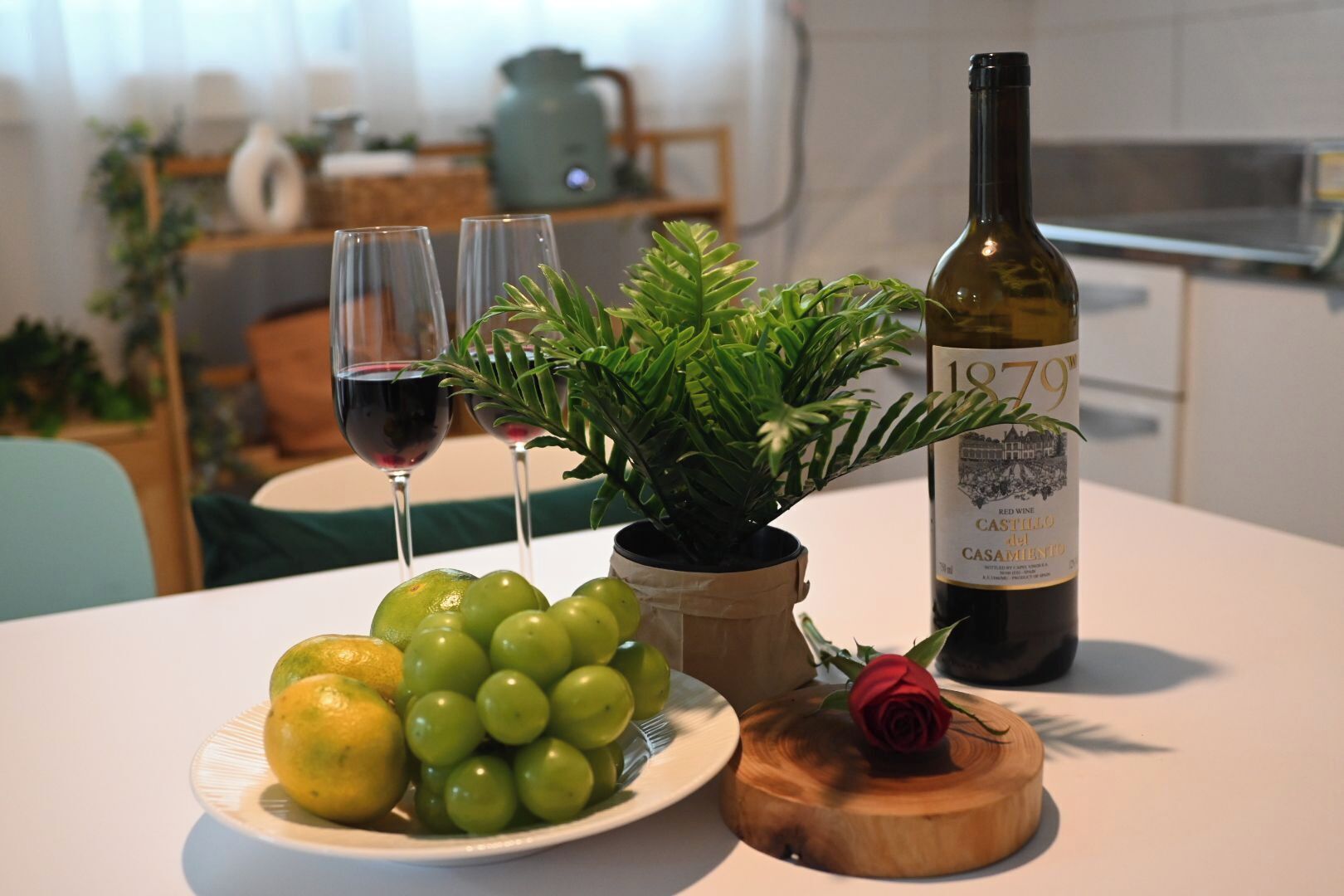 Une table avec une bouteille de vin rouge, une plante en pot, une assiette de raisins et un citron, et une rose posée sur un sous-verre en bois.