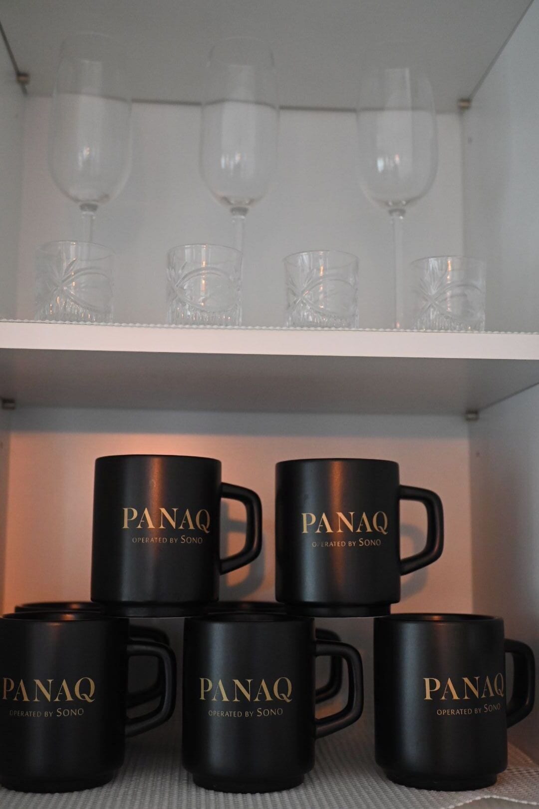 A shelf with black mugs labeled "PANAQ OPERATED BY SONO" and clear glassware above.