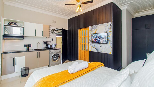 Habitación de hotel pequeña con cama, televisor, ventilador de techo, kitchenette y lavadora.