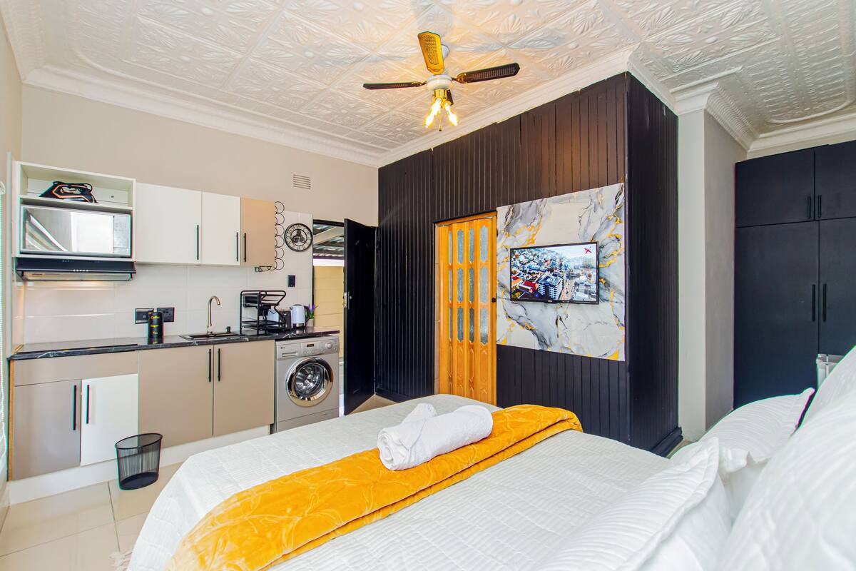 A compact hotel room with a bed, a TV, a ceiling fan, a kitchenette, and a washing machine.