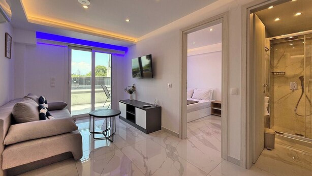 A modern living room with a sofa, a small table, a TV, and a sliding glass door leading to a balcony. - Deluxe One-Bedroom Suite No. 4 (Ground Floor) (Paralia)