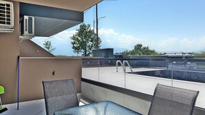 A balcony with a glass table and chairs, air conditioning units, and a view of a swimming pool and mountains. - Deluxe One-Bedroom Suite No. 4 (Ground Floor) (Paralia)