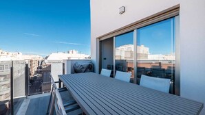Een dakterras met een houten tafel en stoelen, grote ramen met uitzicht op de stad, en een heldere lucht.