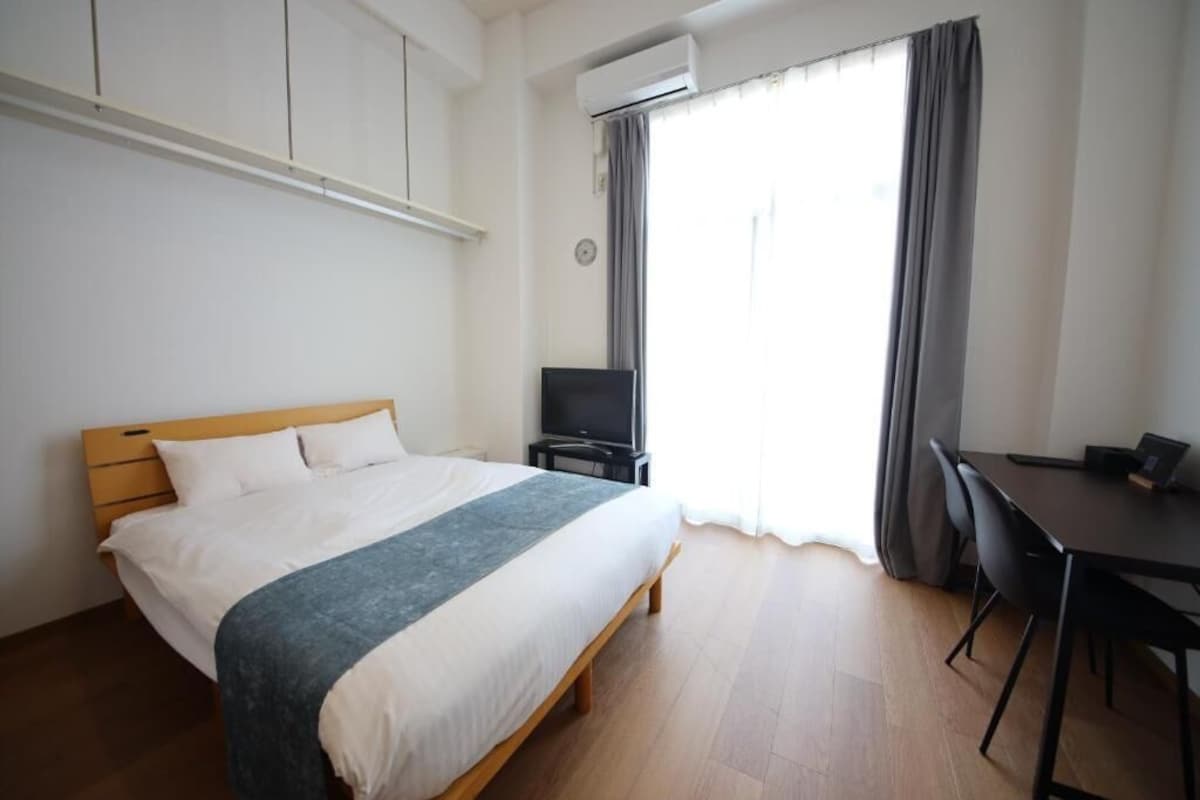 Quarto de hotel com cama, escrivaninha, televisão e janela com cortinas.