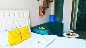 Una habitación de hotel con una cama de cabecera capitonada, un pequeño sofá azul verdoso, una mesita redonda y un dispensador de agua azul.
