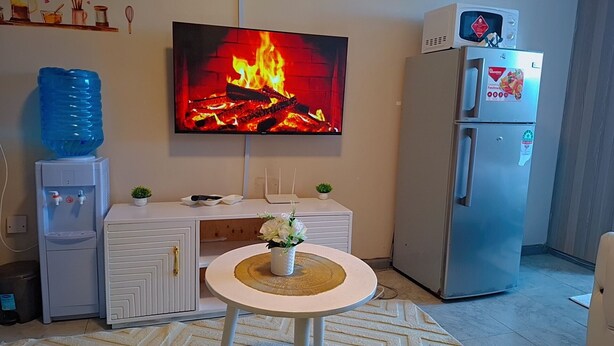 Una sala de estar con televisión de pantalla plana mostrando una chimenea, un dispensador de agua, un refrigerador y una mesita con un jarrón de flores.