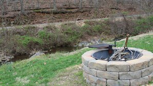 A stone fire pit with a metal grate, surrounded by a grassy area with a stream and trees in the background. - Rural Cabin along Swift Run River, ten minutes from Shenandoah National Park. (Stanardsville)