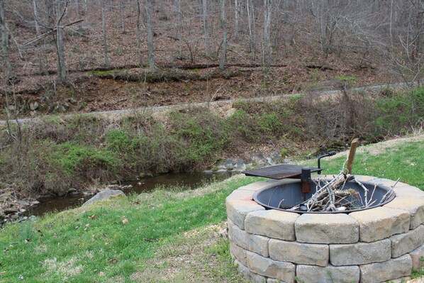 Un hogar de piedra con una parrilla de metal, rodeado de un espacio cubierto de césped, con un arroyo y árboles al fondo.