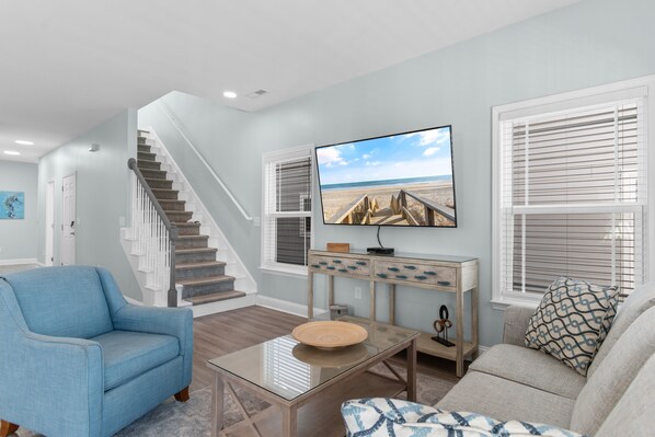 Un salon avec un escalier, un téléviseur fixé au mur et une vue sur la plage par la fenêtre.