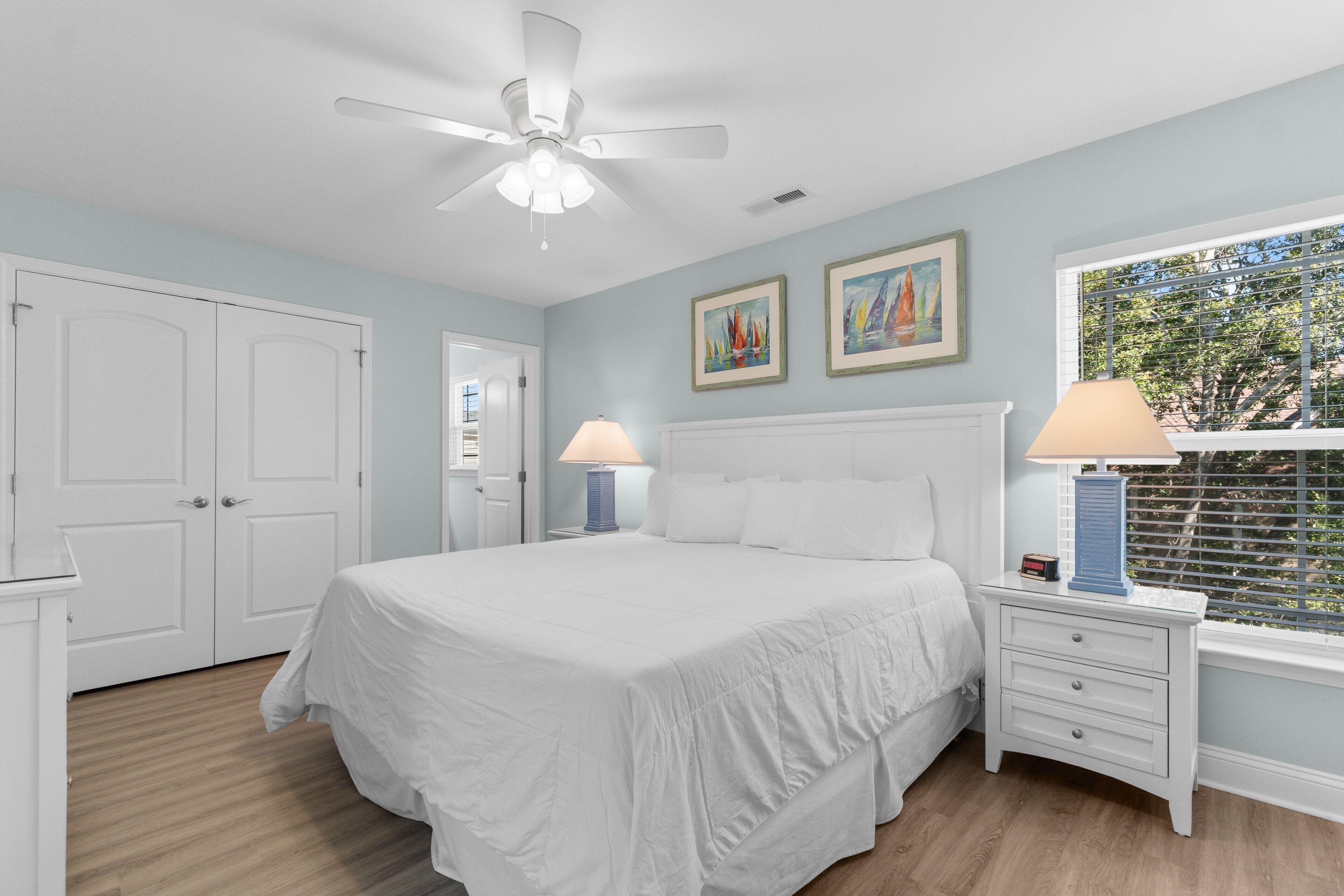 Une chambre à coucher avec un grand lit, deux tables de chevet avec des lampes, un ventilateur de plafond et deux tableaux encadrés accrochés au mur.