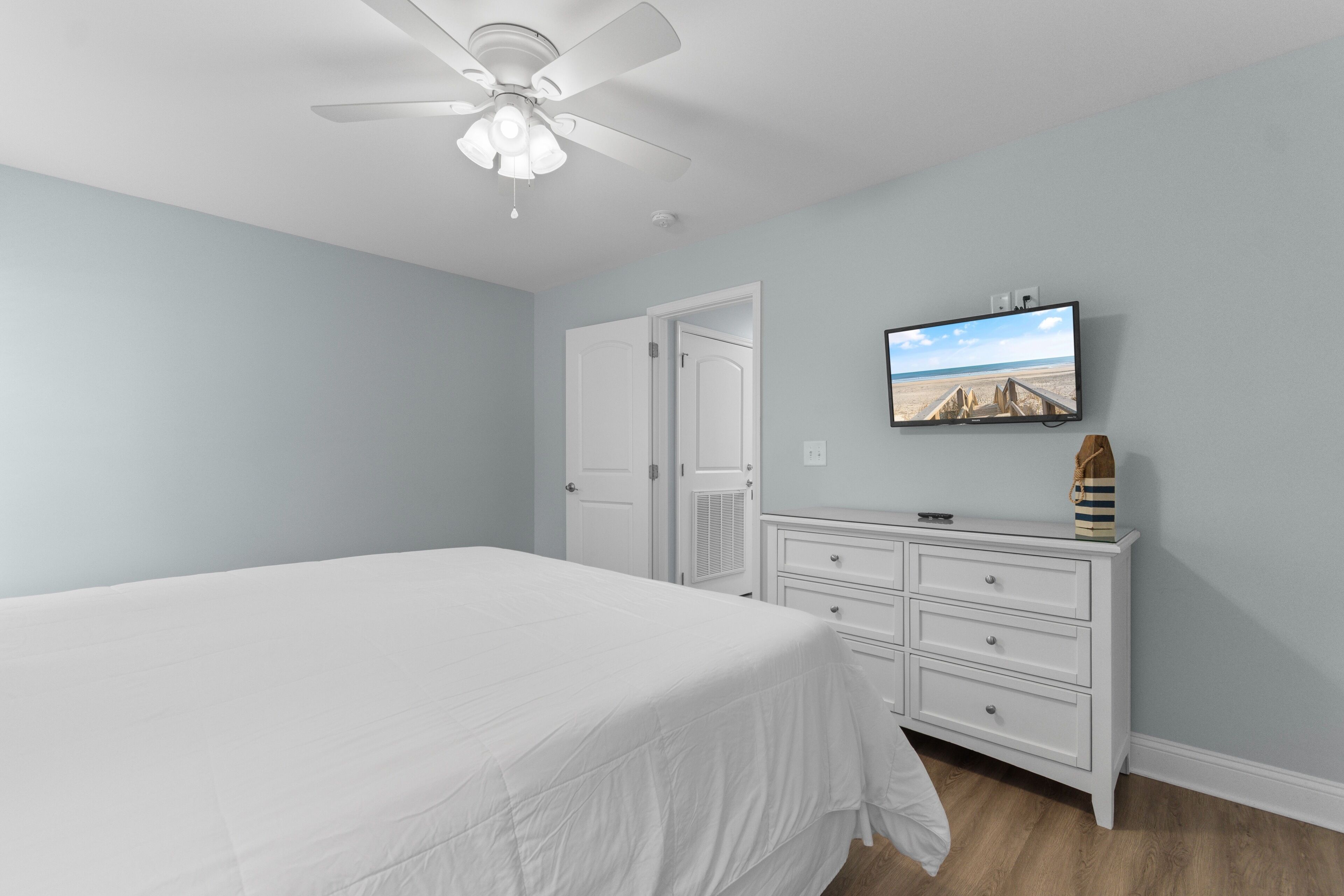 A bedroom with a bed, a dresser, a television on the wall, and a ceiling fan.