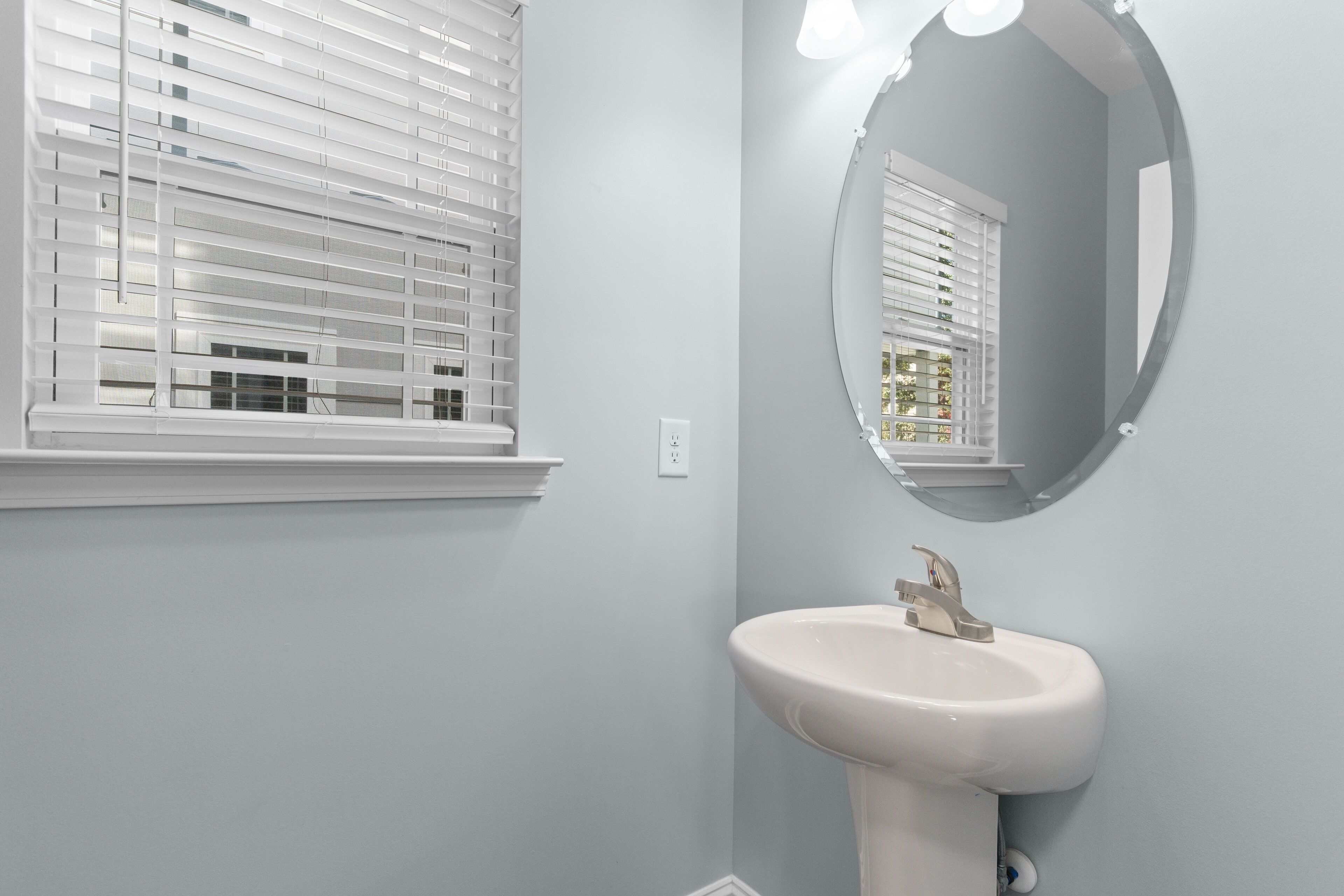A bathroom with a white sink, a round mirror, and white window blinds.