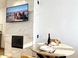 A dining area with a television mounted on the wall, a fireplace, and a table set for two with wine glasses and a bottle.