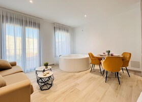 A modern living room with a beige sofa, a round coffee table, a dining table with yellow chairs, and a large white bathtub.