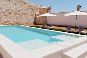 Una piscina rectangular con agua azul cristalina, rodeada por un muro de piedra y una valla blanca. Hay dos sombrillas blancas que dan sombra sobre dos tumbonas.