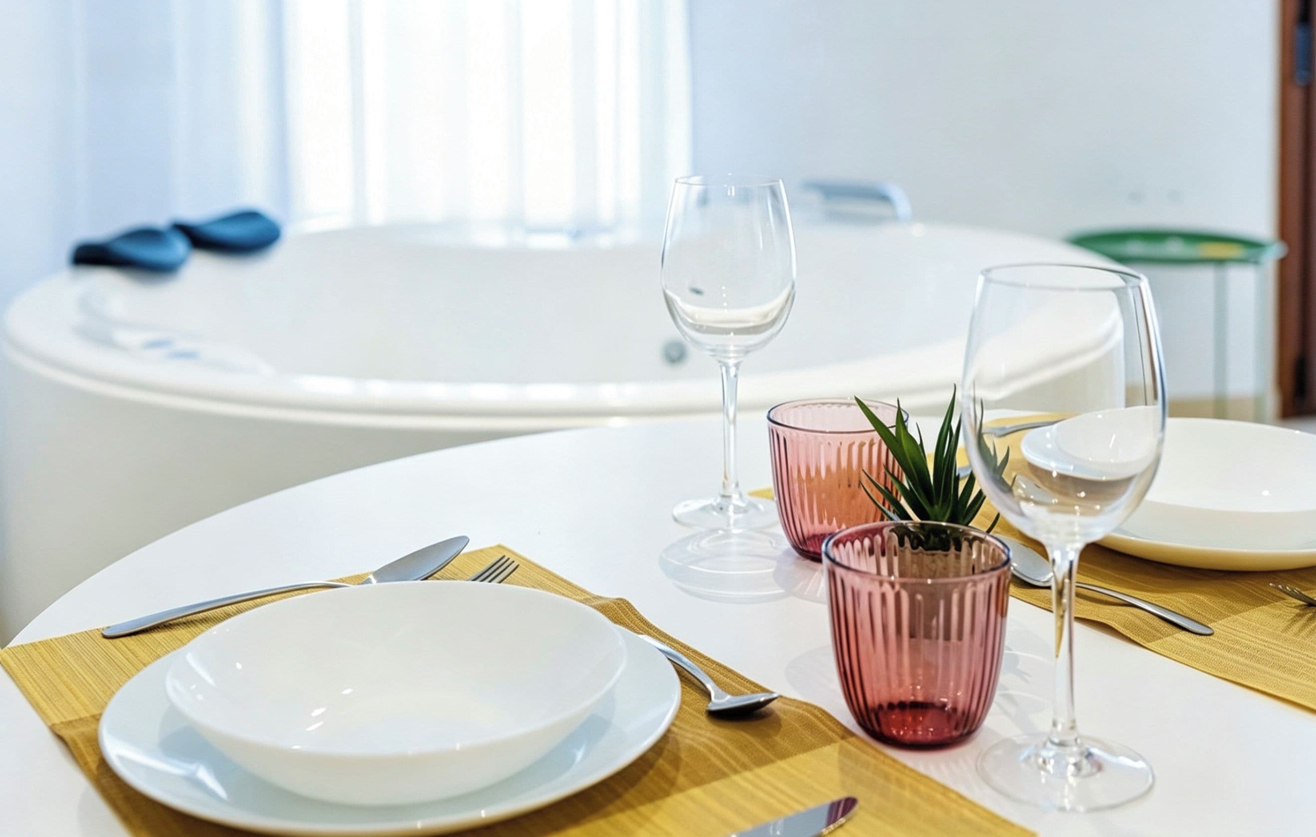 A dining table set with plates, cutlery, and glasses, with a bathtub and a window in the background.