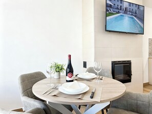 Mesa de comedor servida con platos, cubiertos, una botella de vino y vasos, con un televisor que muestra vista a una piscina.