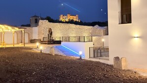 A courtyard with a stone wall, a small building with a window, and a castle in the background. - Complejo La Serrería (Belmonte)
