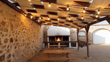 Un patio exterior con muro de piedra, banco de madera, fogata y luces colgantes.