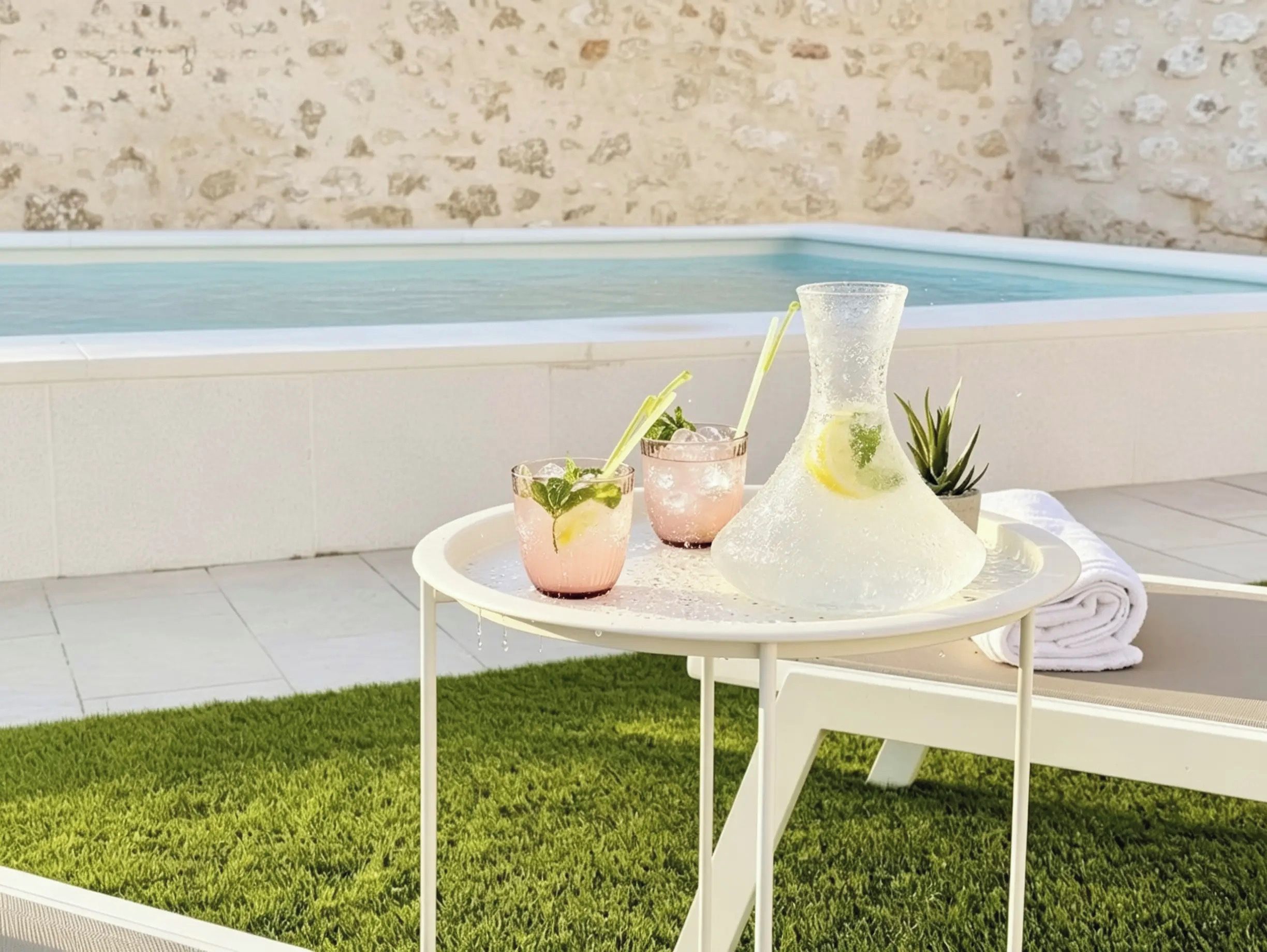 Escena junto a la piscina con una mesa que sostiene una jofaina y dos vasos de una bebida rosada, una planta pequeña en maceta y una toalla enrollada.