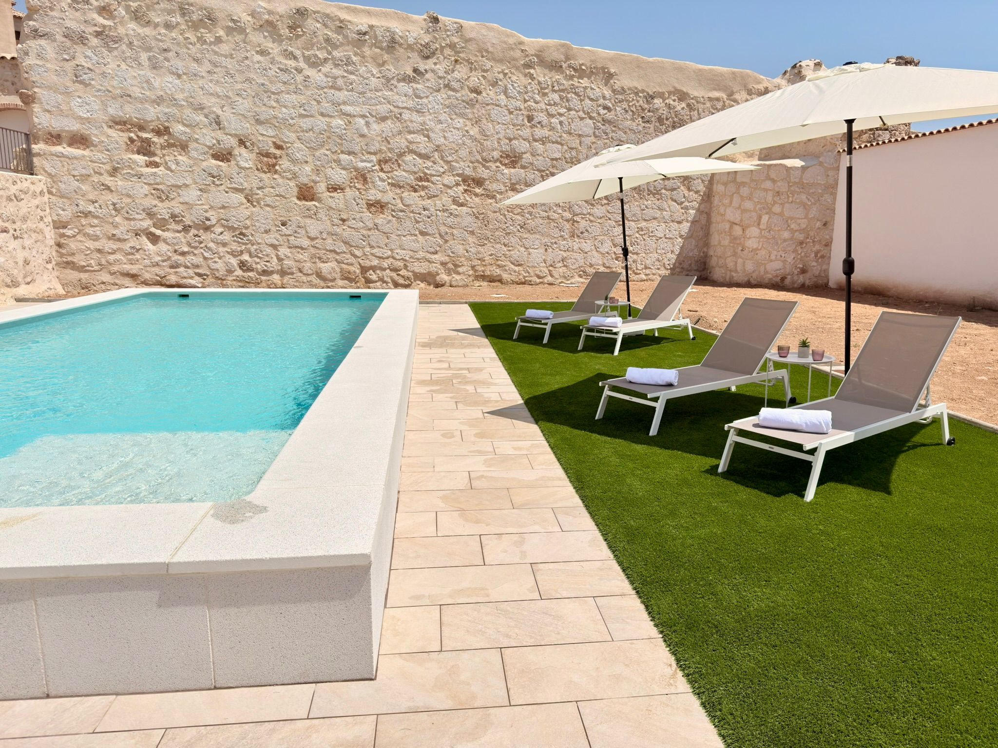 Una piscina con pared de piedra de fondo, rodeada por un patio con césped artificial y sillones de descanso.