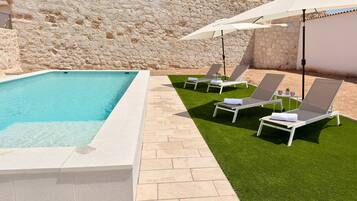 Una piscina con pared de piedra de fondo, rodeada por un patio con césped artificial y sillones de descanso.