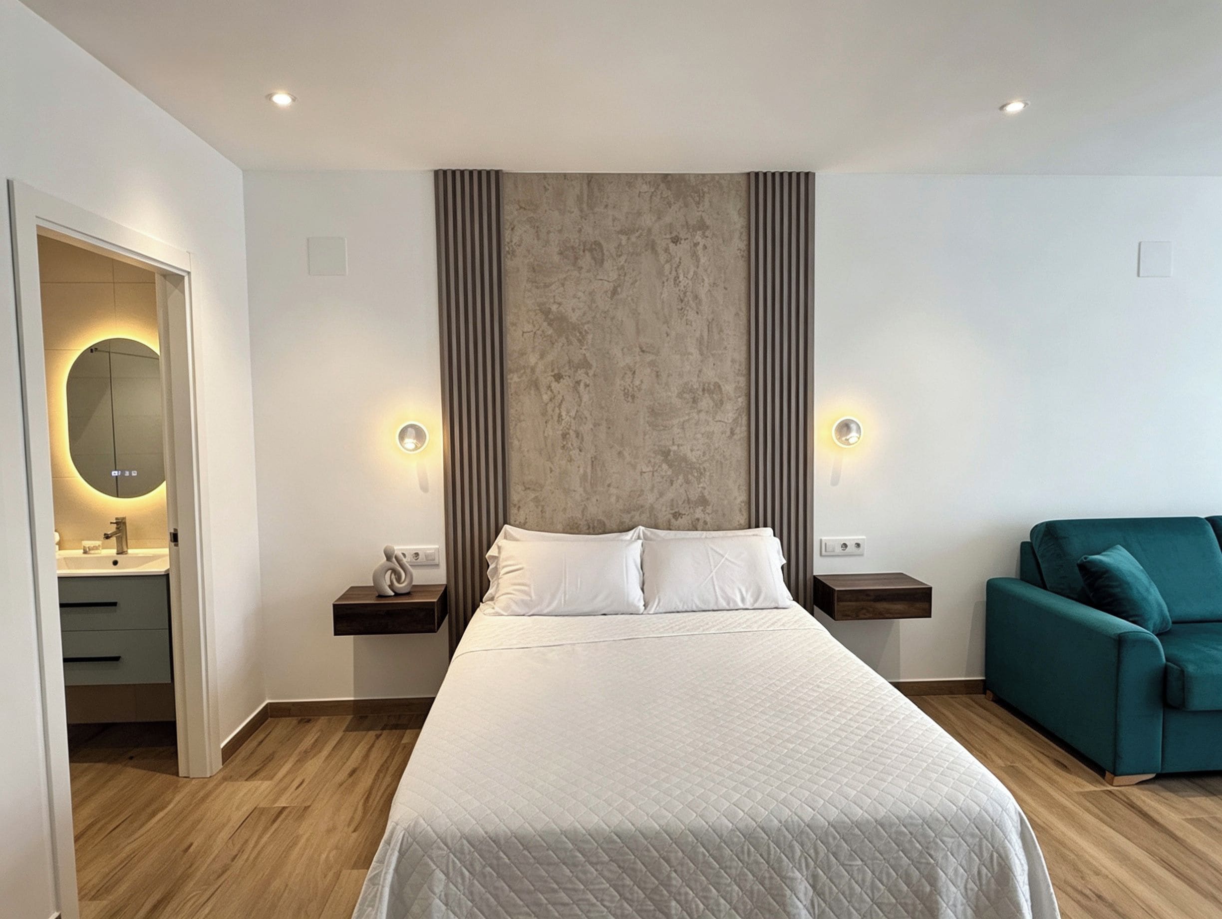 Una habitación de hotel moderna con una cama grande, mesitas de noche, un sofá verde y un baño con espejo y lavamanos.