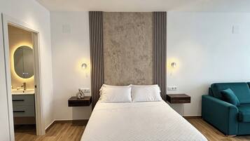 Una habitación de hotel moderna con una cama grande, mesitas de noche, un sofá verde y un baño con espejo y lavamanos.