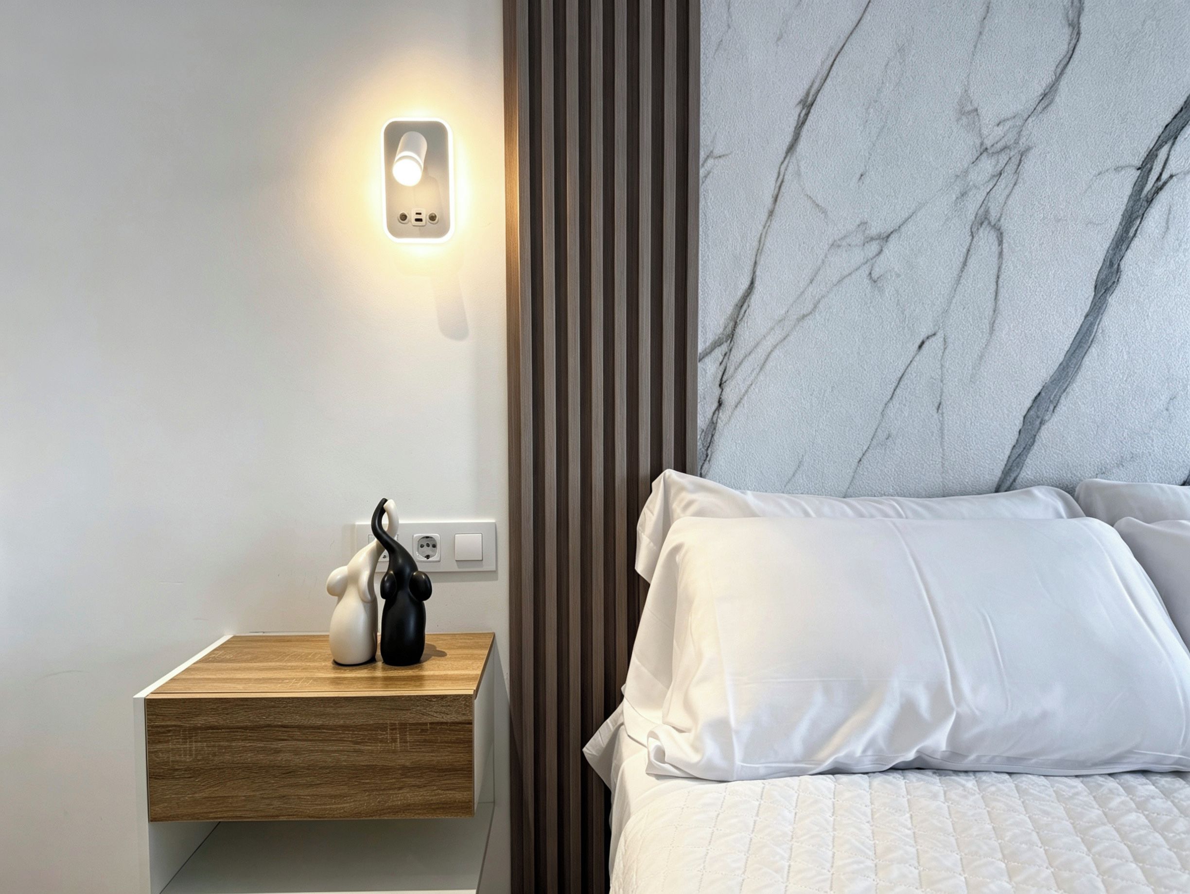 A modern bedroom with a wooden bedside table, a nightstand with a wall-mounted light, a marble-patterned headboard, and white bedding.