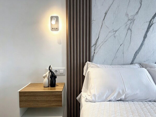 A modern bedroom with a wooden bedside table, a nightstand with a wall-mounted light, a marble-patterned headboard, and white bedding. - Complejo La Serrería (Belmonte)