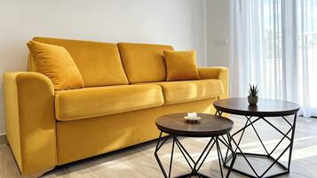 Un sofá amarillo con tres cojines, una mesita redonda con una vela y una planta en maceta.