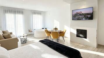 A modern hotel room with a bed, a seating area with a table and chairs, a jacuzzi, and a TV mounted on the wall.
